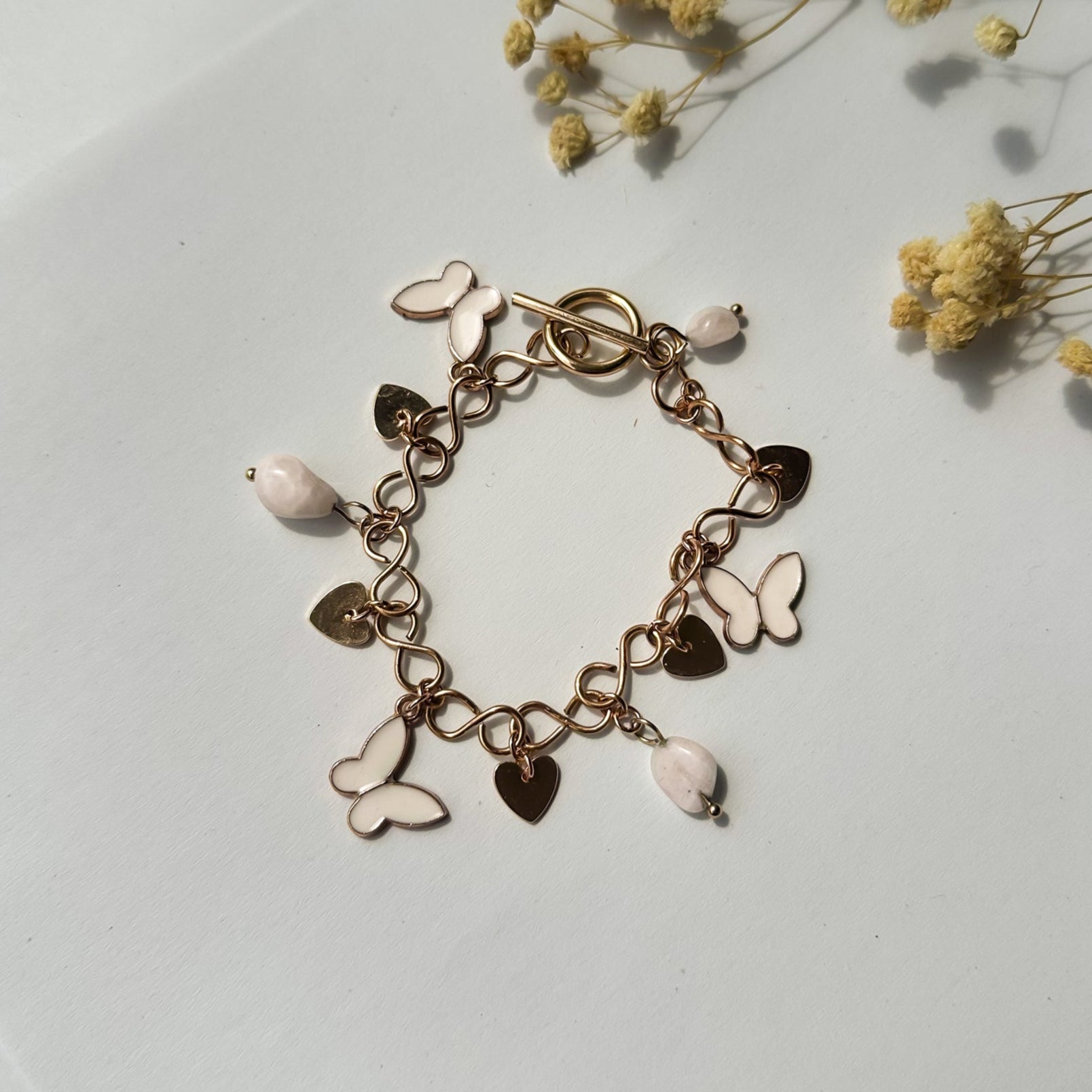 Flach liegendes Goldarmband mit weißen Schmetterlingen und Herzen, dekoriert mit Trockenblumen – romantischer Schmuckstil.