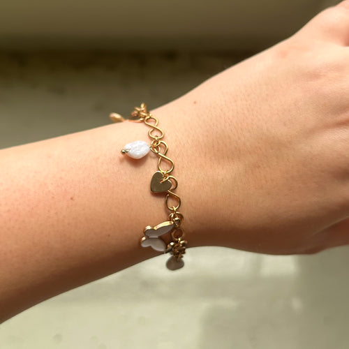Goldarmband mit weißen Schmetterlings-, Mondstein- und Herzanhängern am Handgelenk einer Frau – zarter Modeschmuck im Sommerlook.