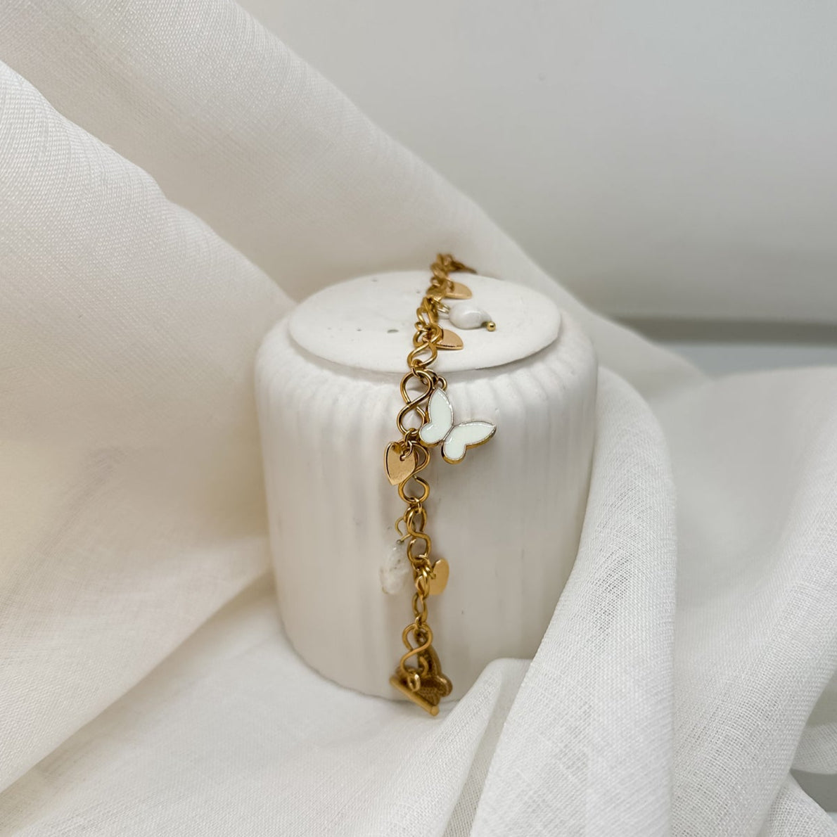 Seitliche Ansicht eines goldfarbenen Armbands mit weißen Schmetterlingsanhängern auf hellem Stoff – femininer Armschmuck