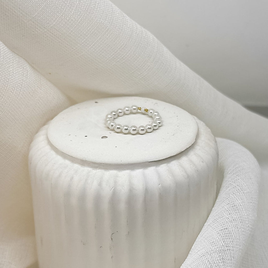 Zarter Perlenring aus weißen Perlen mit einzelnen goldfarbenen Details, dekorativ platziert auf weißem Stoff im minimalistischen Stil.