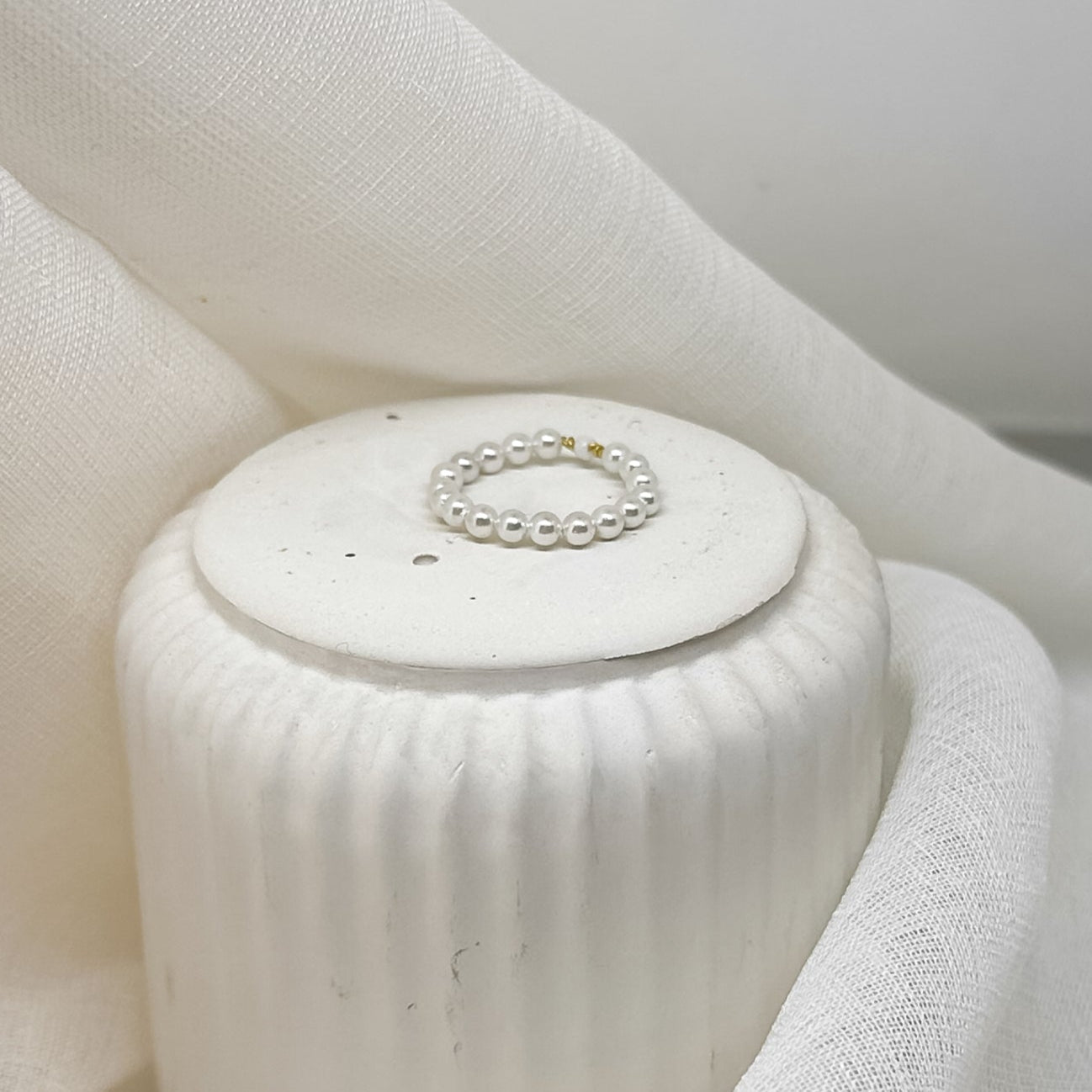 Zarter Perlenring aus weißen Perlen mit einzelnen goldfarbenen Details, dekorativ platziert auf weißem Stoff im minimalistischen Stil.