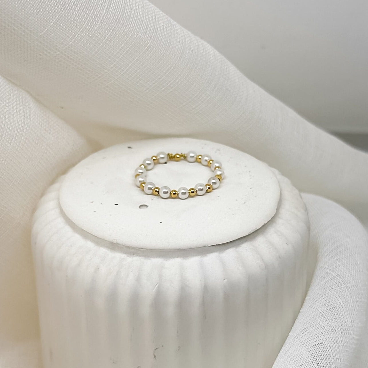 Feiner Perlenring aus weißen Perlen und goldfarbenen Zwischenelementen, stilvoll arrangiert auf weißem Stoff im minimalistischen, eleganten Look