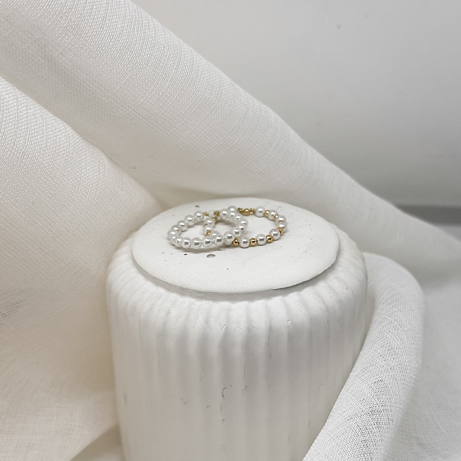 Zwei zarte Perlenringe aus weißen Perlen mit goldfarbenen Details, dekorativ arrangiert auf weißem Stoff, minimalistischer Schmuck im eleganten Stil.